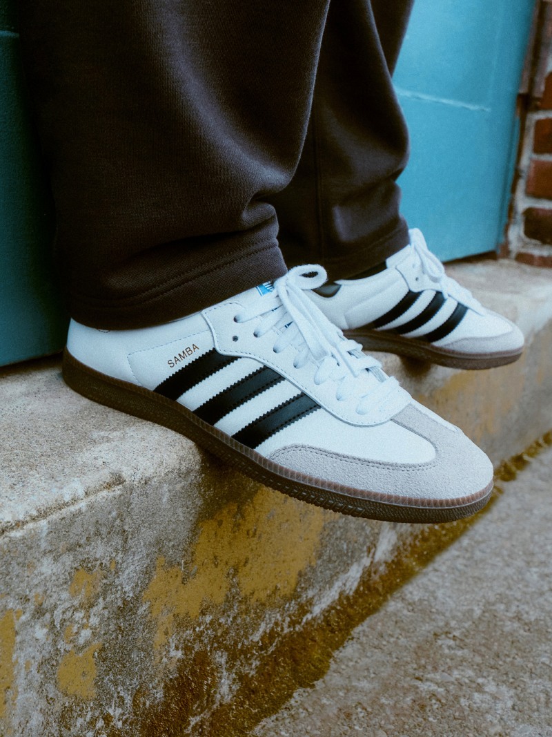 A pair of white and black Adidas sneakers rests on a worn, yellow-tinted concrete surface, with a dark background visible behind them.
