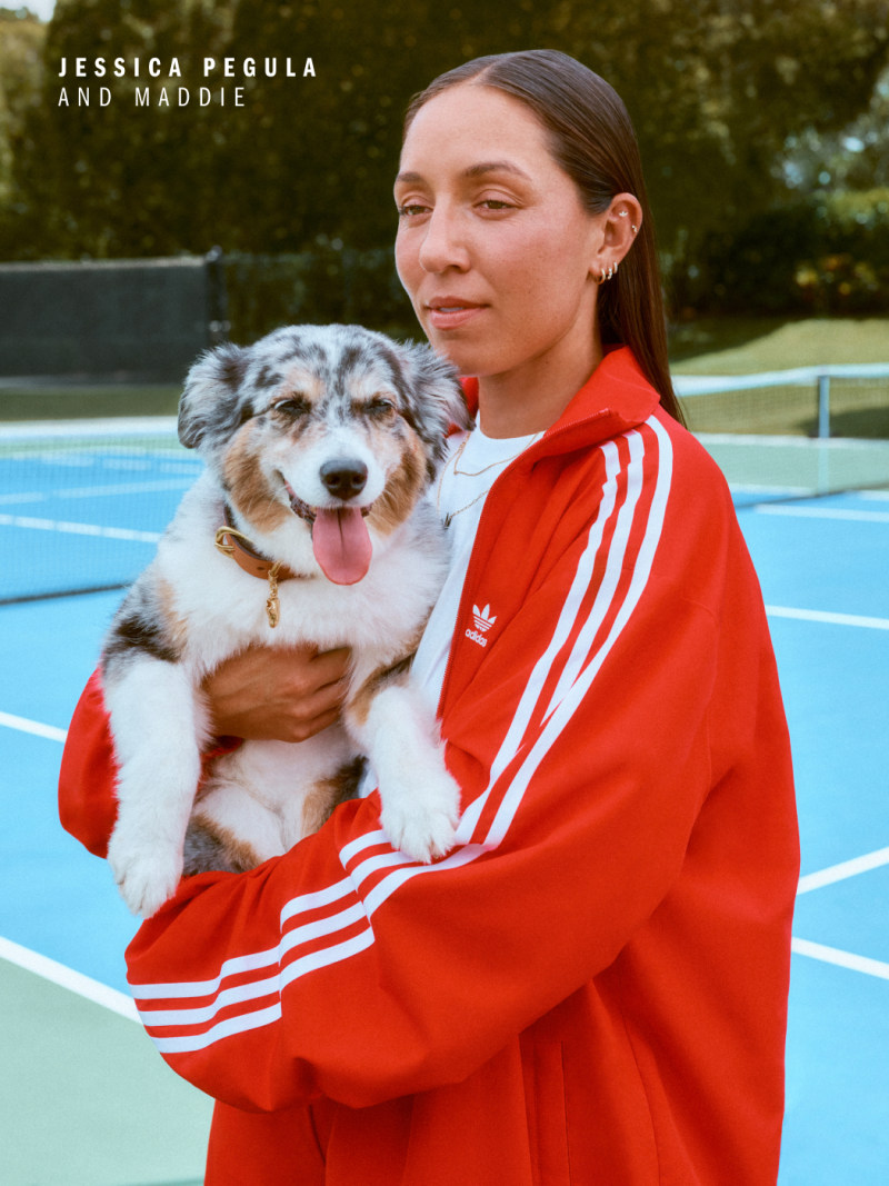 A young woman in a red Adidas tracksuit is holding a smiling Australian Shepherd dog on a tennis court.