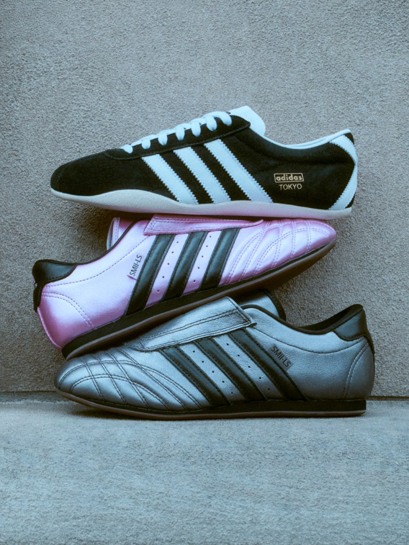 The image shows a pair of Adidas soccer cleats in black and white, with a pink pair of cleats in the background, all resting on a concrete surface.
