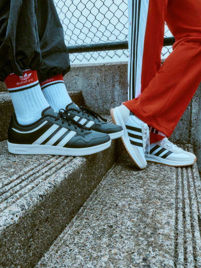 The image shows a pair of Adidas sneakers, one black and one white, on a concrete surface with a metal fence in the background.