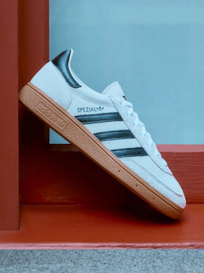 A white and blue Adidas Spezial sneaker rests on a red and blue background, with a plain blue wall visible in the background.