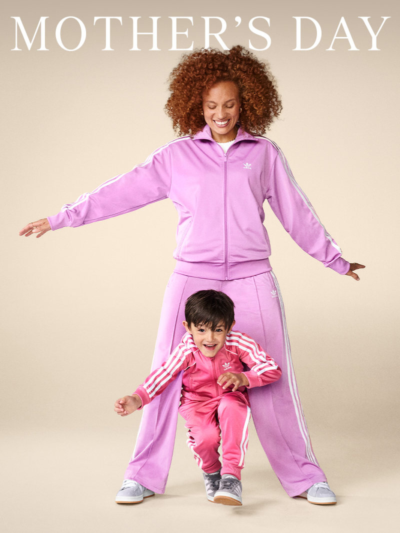 A young woman with curly hair wearing a pink tracksuit is standing with her arms outstretched, while a young boy in a pink outfit is sitting on the floor in front of her.