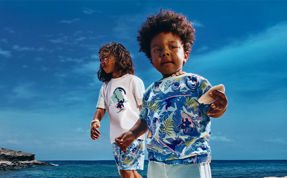Two young children, a boy and a girl, stand together on a beach with a vibrant blue ocean and sky in the background.