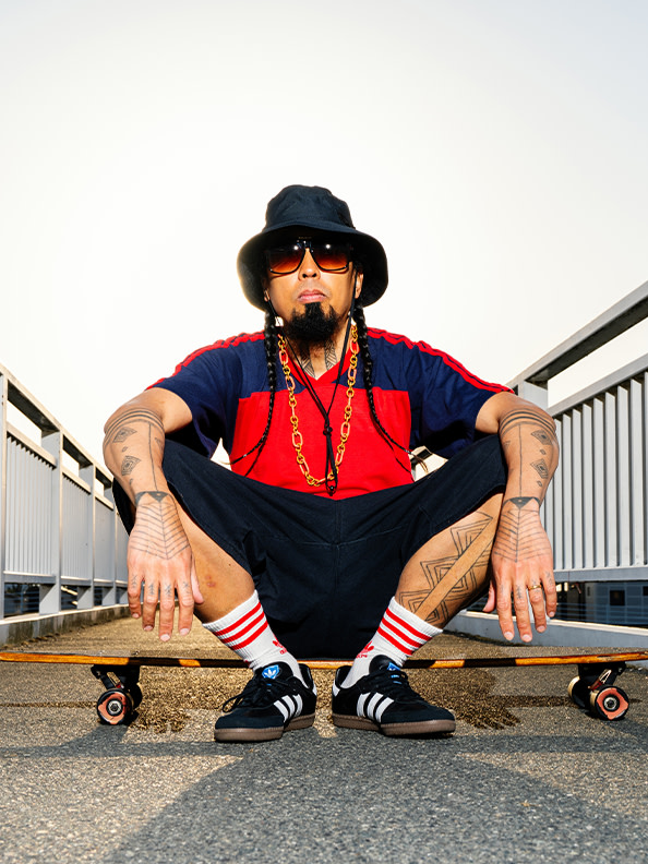 A man with a beard and sunglasses, wearing a red and blue shirt, sits on a skateboard in front of a row of storage containers.