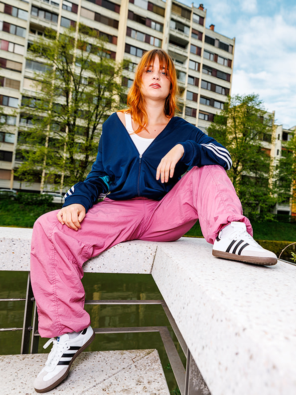 A young woman with red hair sits on a concrete ledge, wearing a navy blue top and bright pink pants, with apartment buildings visible in the background.