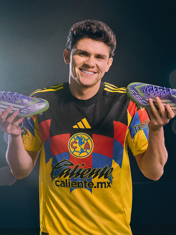 A smiling young man wearing a yellow and red soccer jersey holds two soccer cleats in his hands against a dark background.