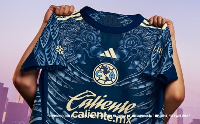 A navy blue and gold soccer jersey with a distinctive eagle design is being held up against a pink and purple urban background.