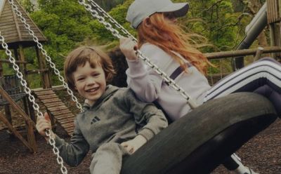 Two children, a boy and a girl, are playing on a swing set in a wooded area with lush greenery surrounding them.