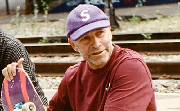 An older man wearing a purple hat and a red shirt stands in a wooded area with a wooden structure in the background.