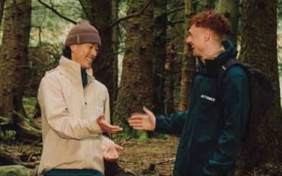 Two individuals, a man and a woman, are standing in a lush, forested environment, engaged in conversation. The background is filled with tall trees and dense vegetation, creating a serene and natural setting.