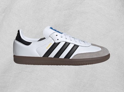 A pair of white and black Adidas sneakers with a gum sole is displayed against a light gray background.