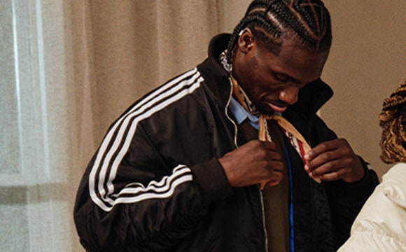 The image shows a person wearing a black jacket with white stripes, standing in what appears to be a room with curtains in the background.