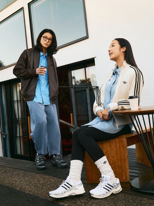 Two young women, one wearing a brown jacket and the other in an Adidas tracksuit, are sitting on a bench in front of a glass building.
