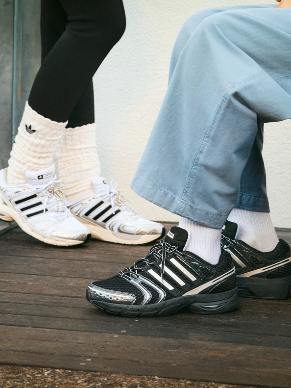 The image shows a pair of black and white athletic shoes, likely Adidas brand, worn by two individuals standing on a wooden surface.