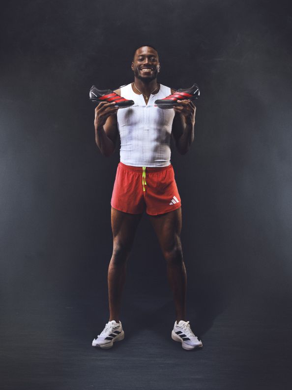 A muscular man in a white tank top and red shorts stands against a dark background, his arms outstretched and a determined expression on his face.