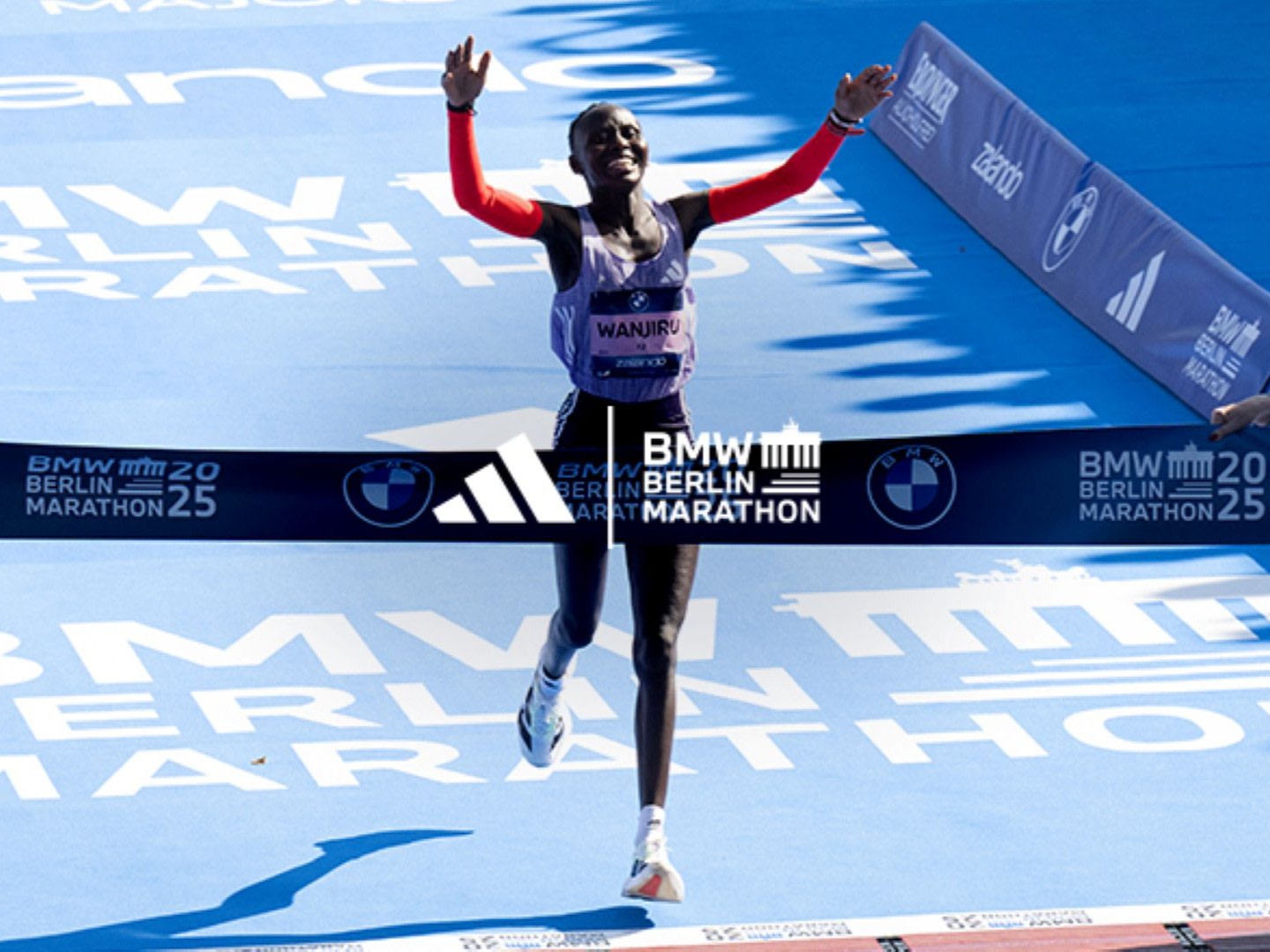 A female runner with arms raised in celebration crosses the finish line of the BMW Berlin Marathon, with the event's branding and banners visible in the background.