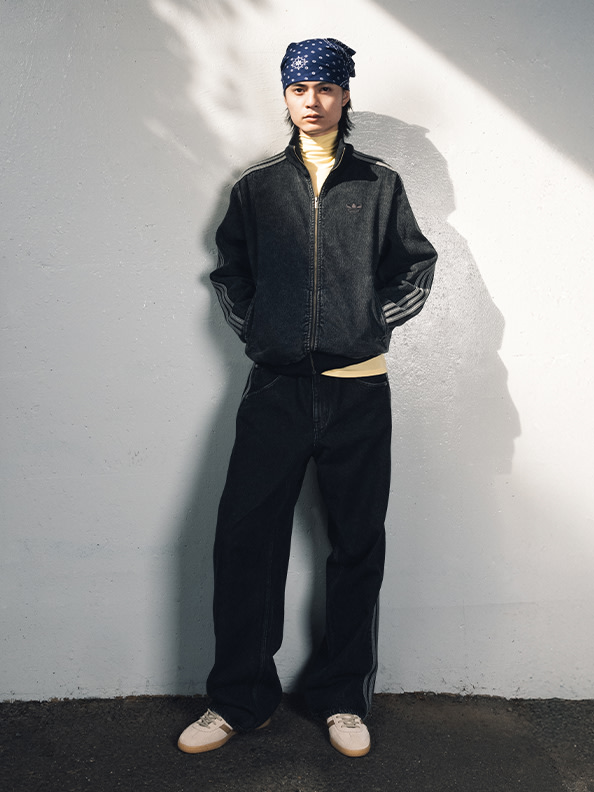 A person wearing a black jacket, black pants, and a patterned headband stands in front of a plain white wall, casting a shadow on the ground.