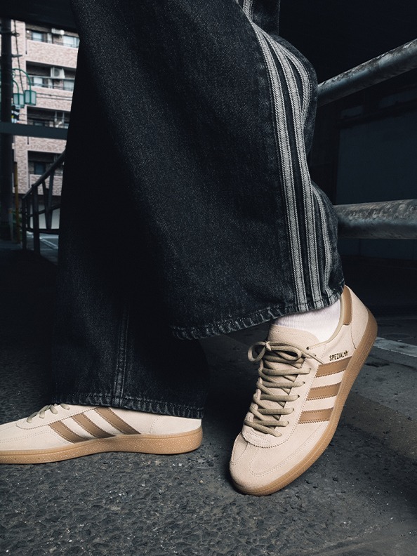 The image shows a pair of beige Adidas sneakers on a dark background, with a building visible in the background.