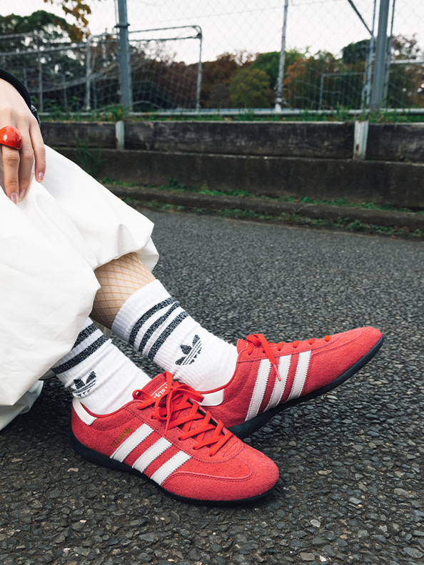 The image shows a person's lower body wearing a white sweater, black and white striped socks, and bright red Adidas sneakers, standing on a paved surface with a fence and greenery in the background.