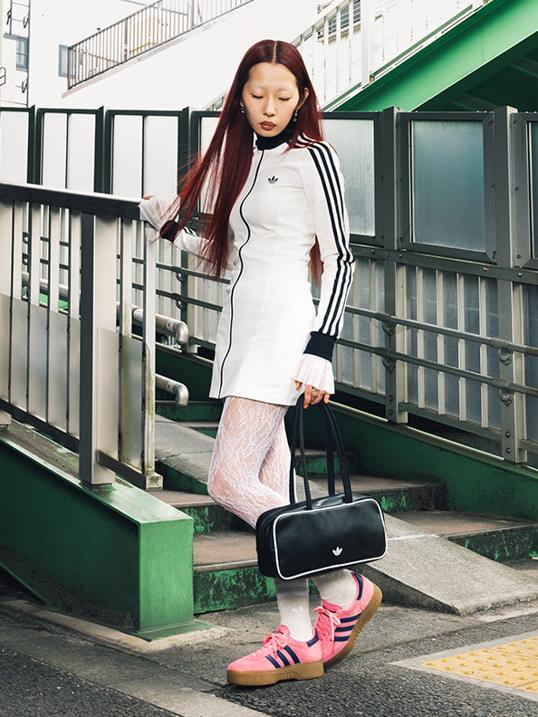 A young woman with long brown hair is standing on a set of stairs, wearing a white jacket with stripes, pink sneakers, and carrying a black bag.