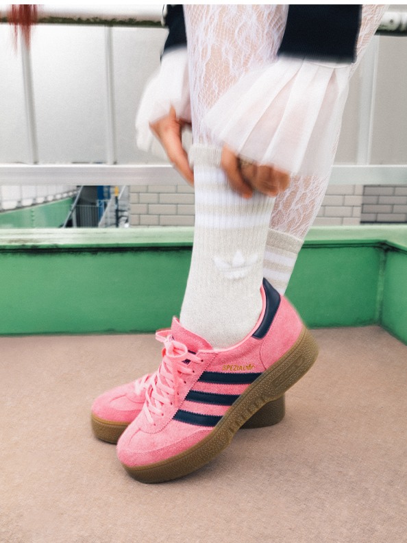 The image shows a person's lower body wearing white pants and pink sneakers with navy blue stripes, standing on a tiled floor with a green wall in the background.