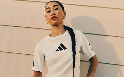 A person wearing a white Adidas t-shirt stands in front of a wooden wall, with a serious expression on their face.