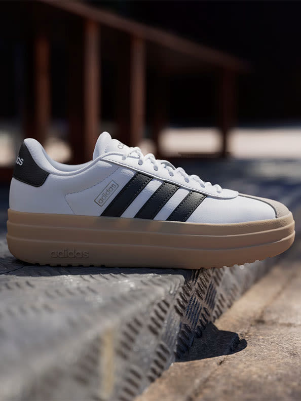 A pair of white and black Adidas sneakers rests on a wooden surface, with a blurred background suggesting an indoor setting.