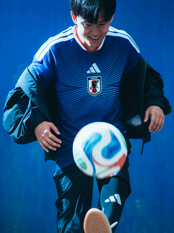 A young soccer player in a blue and white jersey stands against a vibrant blue background, holding a soccer ball and smiling.