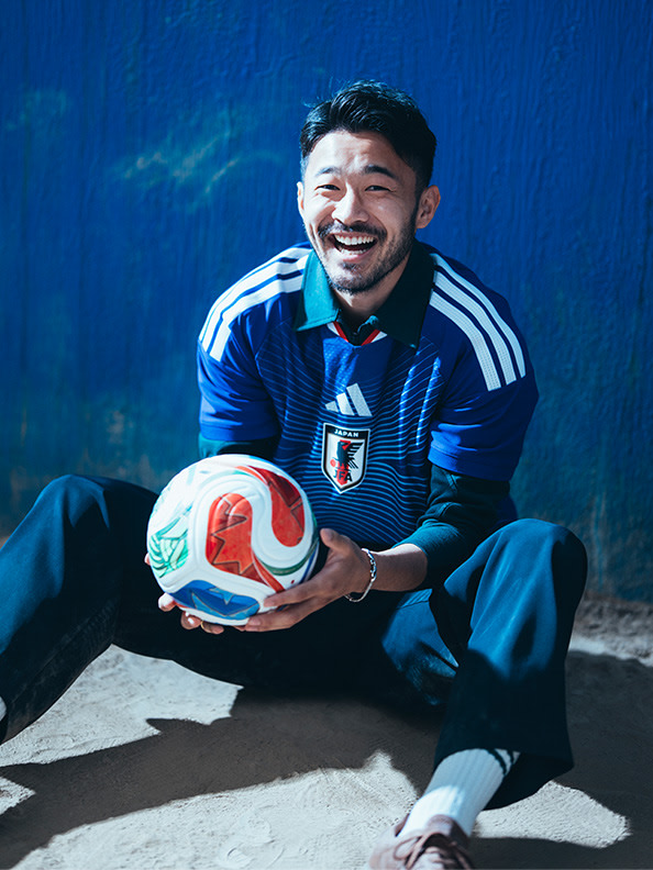 A smiling man in a blue and white sports jersey is sitting on the ground and holding a soccer ball.