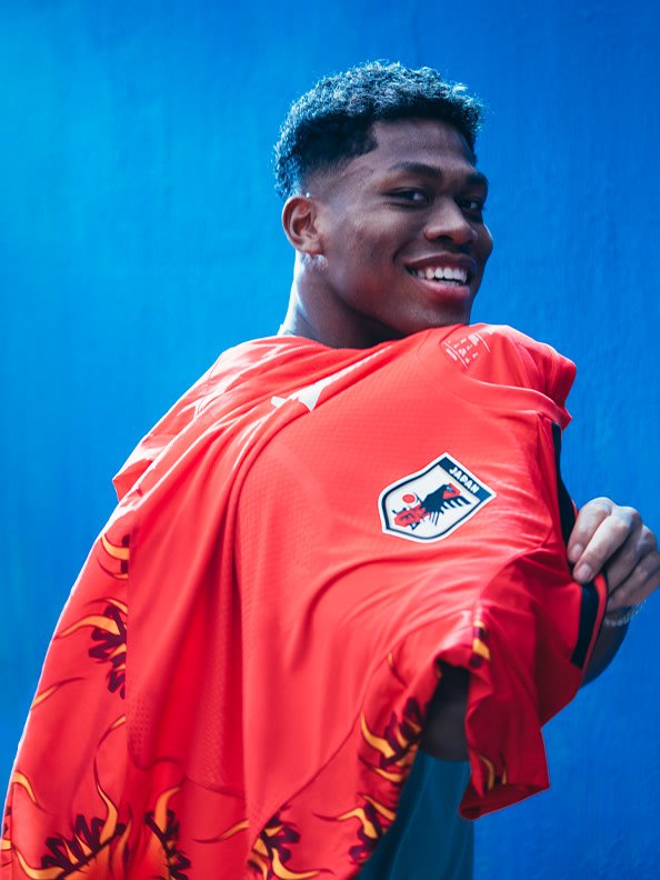 A young man wearing a bright red jersey is smiling against a vibrant blue background.