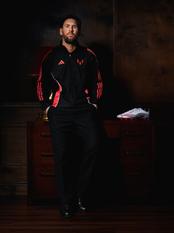 A man in a black and red athletic jacket stands in a dimly lit room, with a pair of sneakers visible on a wooden surface in the foreground.