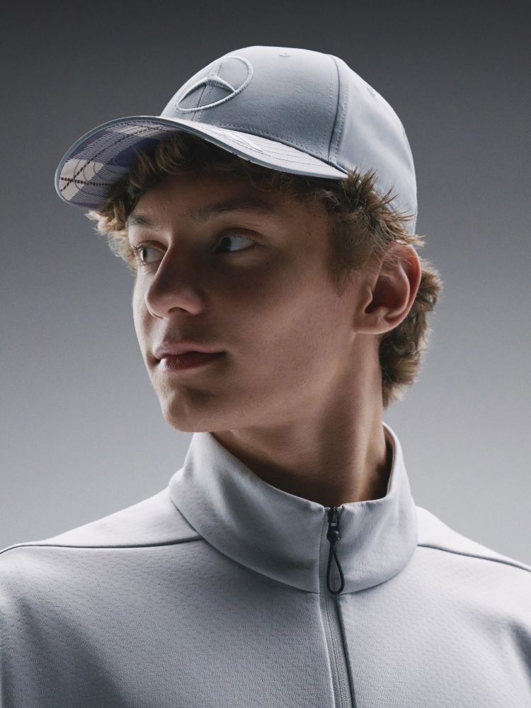A young man wearing a gray athletic jacket and cap, with a serious expression on his face, stands against a plain gray background.
