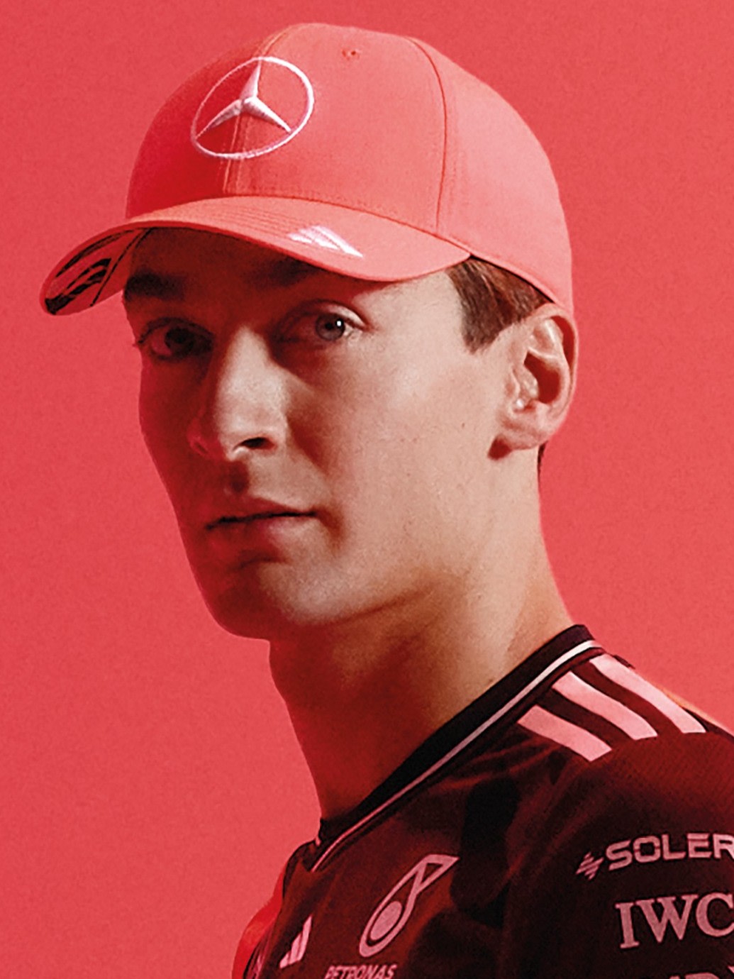 A young man wearing a red baseball cap with a Mercedes-Benz logo and a black and red jersey stands against a vibrant red background.