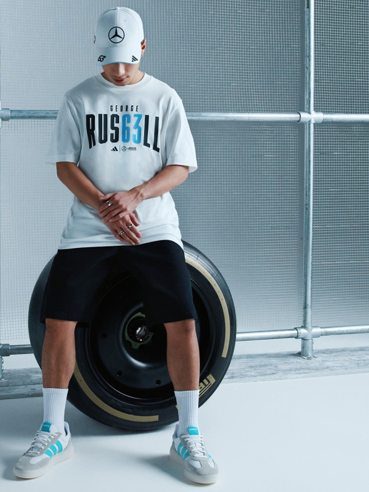 A person wearing a white t-shirt with "George Russell" printed on it, sitting on a large tire-like object against a gray background.