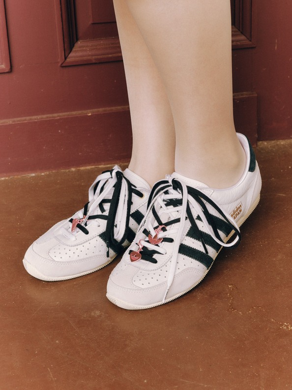 The image shows a pair of white sneakers with black and red accents, resting on a wooden floor in front of a red-painted wall.