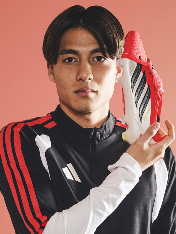 A young man wearing a black and red sports jersey stands against a coral-colored background, holding a red and white soccer ball.