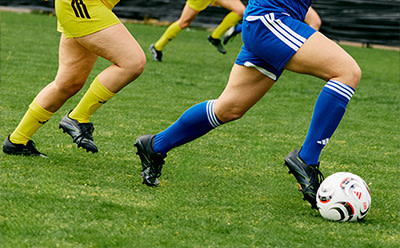 The image shows the lower bodies of two soccer players in action on a grassy field, with one player in a yellow uniform and the other in a blue uniform, both wearing cleats and chasing after a soccer ball.