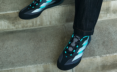The image shows a pair of black and teal-colored athletic shoes with a camouflage-like pattern on the sides, standing in front of a concrete wall.