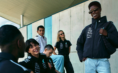 A group of people, some wearing black clothing with a logo, are standing and interacting with each other in what appears to be an indoor setting with a white wall in the background.