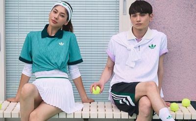 Two individuals, a woman and a man, are standing in front of a white metal shutter backdrop, wearing athletic-style clothing with the Adidas logo visible.