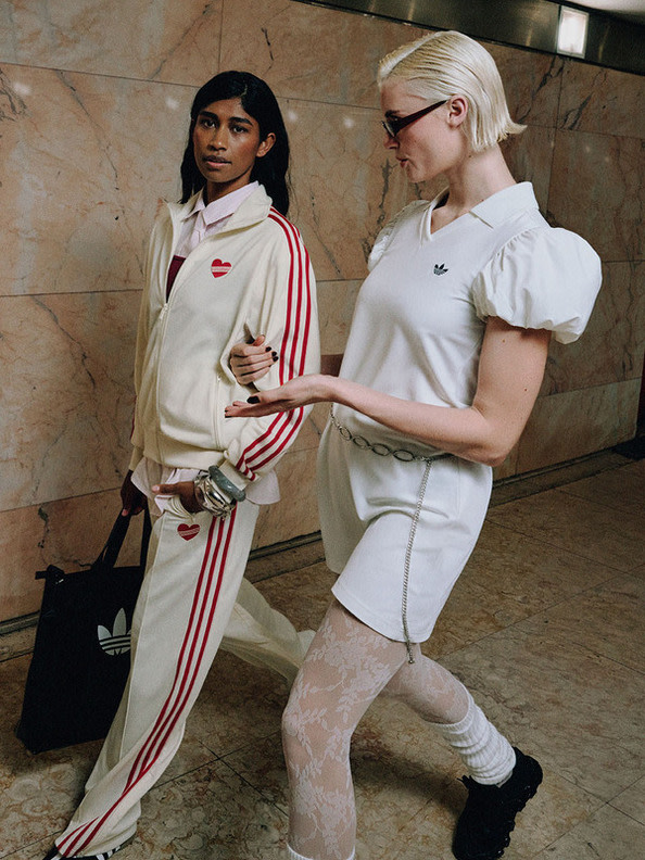 Two young women, one with dark hair and the other with blonde hair, are standing together in a room with a tiled wall in the background.