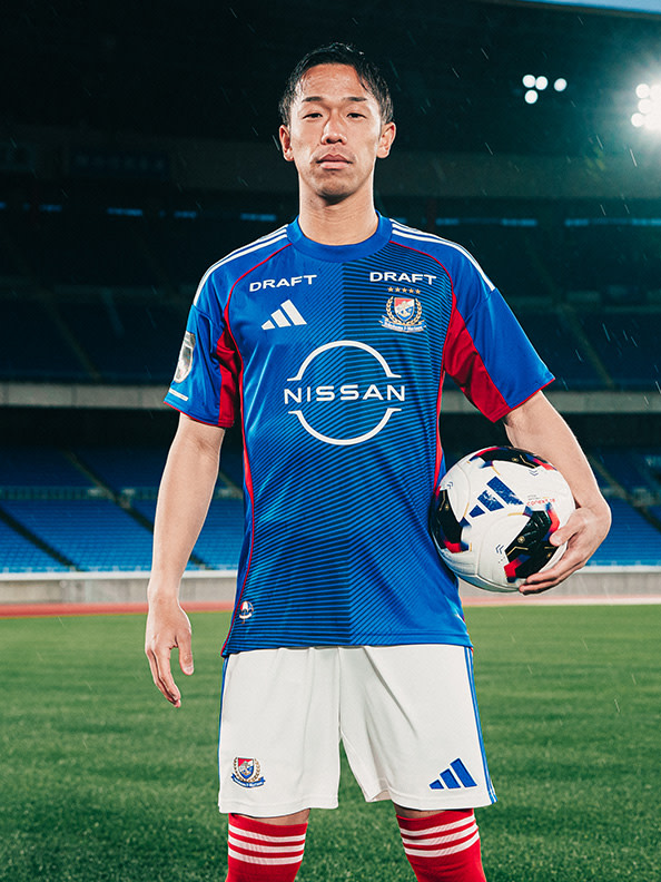 A young man wearing a blue and red soccer jersey stands on a soccer field, holding a soccer ball.