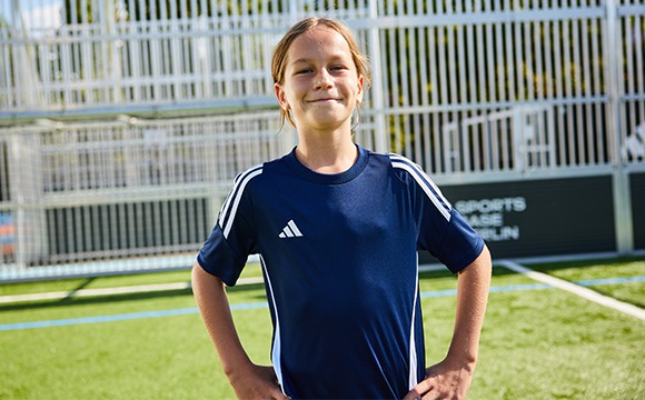 A young person wearing a navy blue sports jersey stands on a grassy field with a chain-link fence in the background.