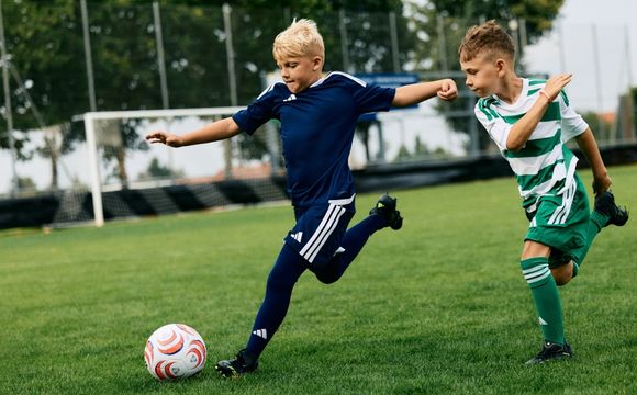 Two young soccer players, one in a navy blue uniform and the other in a green and white striped uniform, are competing for the ball on a grassy soccer field with a goal post visible in the background.