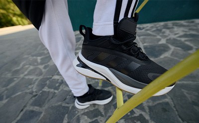 The image shows a pair of black and white athletic shoes on a person's feet, standing on a paved surface with a blurred green background.
