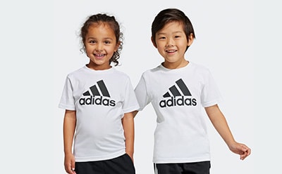 Two young girls wearing white Adidas t-shirts are smiling and standing against a plain background.