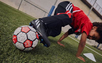A person in a red and black sports uniform is crouched down on a grassy field, with a soccer ball in front of them.