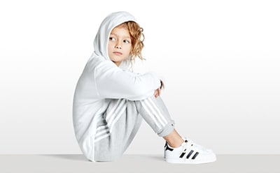 A young child wearing a white hooded garment and Adidas sneakers stands against a plain white background.