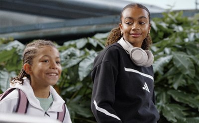 Two young girls, one with curly hair and the other with straight hair, are standing in a lush, green outdoor setting with plants and foliage in the background.