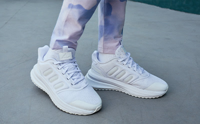 The image shows a pair of white sneakers with a distinctive design, worn by someone wearing tie-dye patterned pants, standing on a concrete surface.
